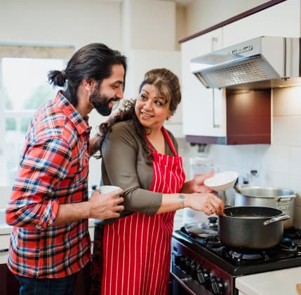 Mature woman is cooking in her kitchen and her son is looking over her shoulder to see what she is making.